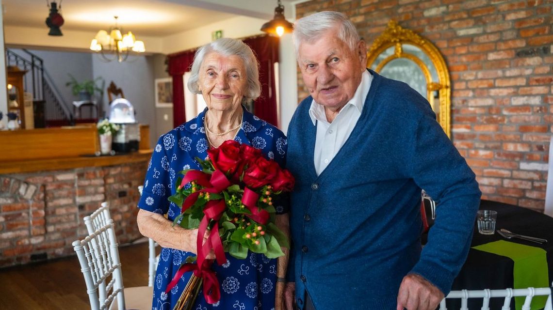 zdjęcie przedstawia portret starszej uśmiechniętej kobiety i mężczyzny, kobieta trzyma czerwone róże