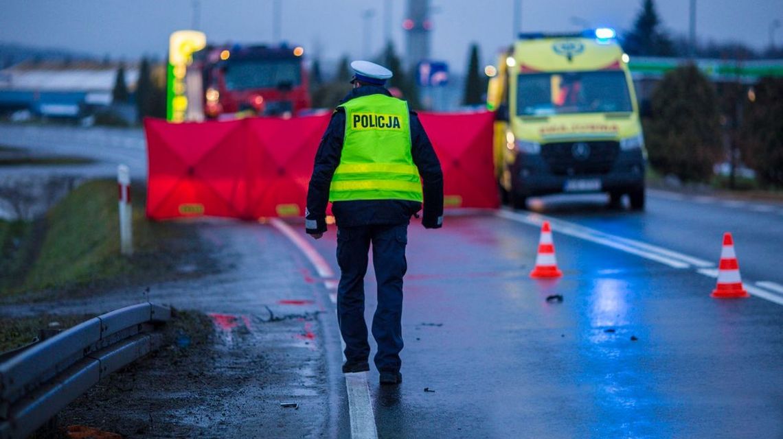 policjant widziany od tył, w tle czerwony parawan, karetka pogotowia i wóz straży pożarnej