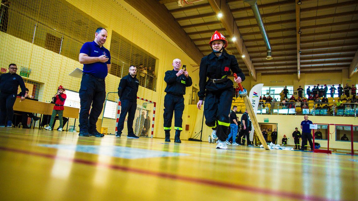 zdjęcie przedstawia dziewczynę strażaczkę biegnącą z wężem podczas zawodów w hali