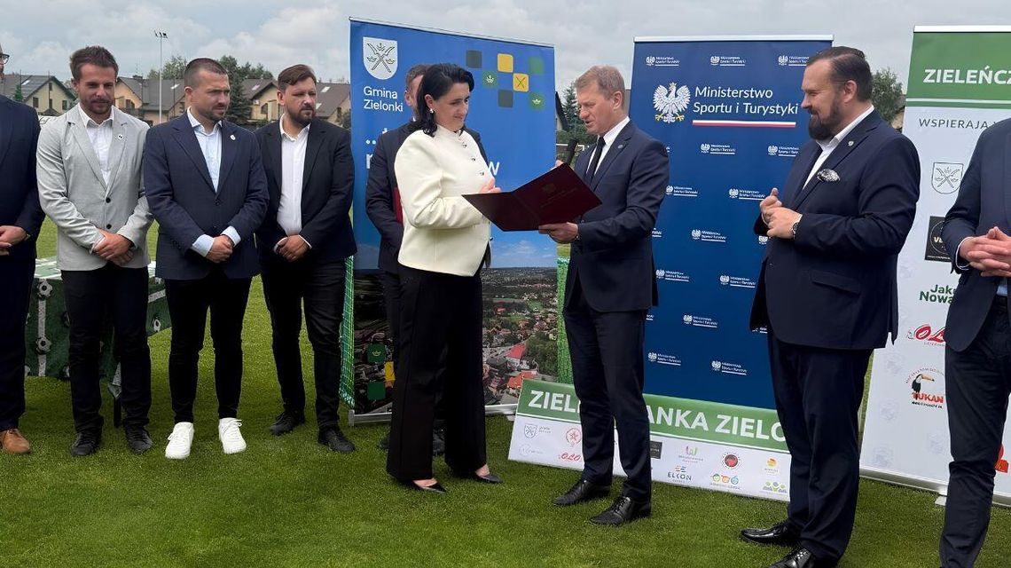 zdjęcie przedstawia grupę osób na boisku sportowym, za nimi banery reklamowe, mężczyzna trzyma przed sobą dokumenty w otwartej teczce
