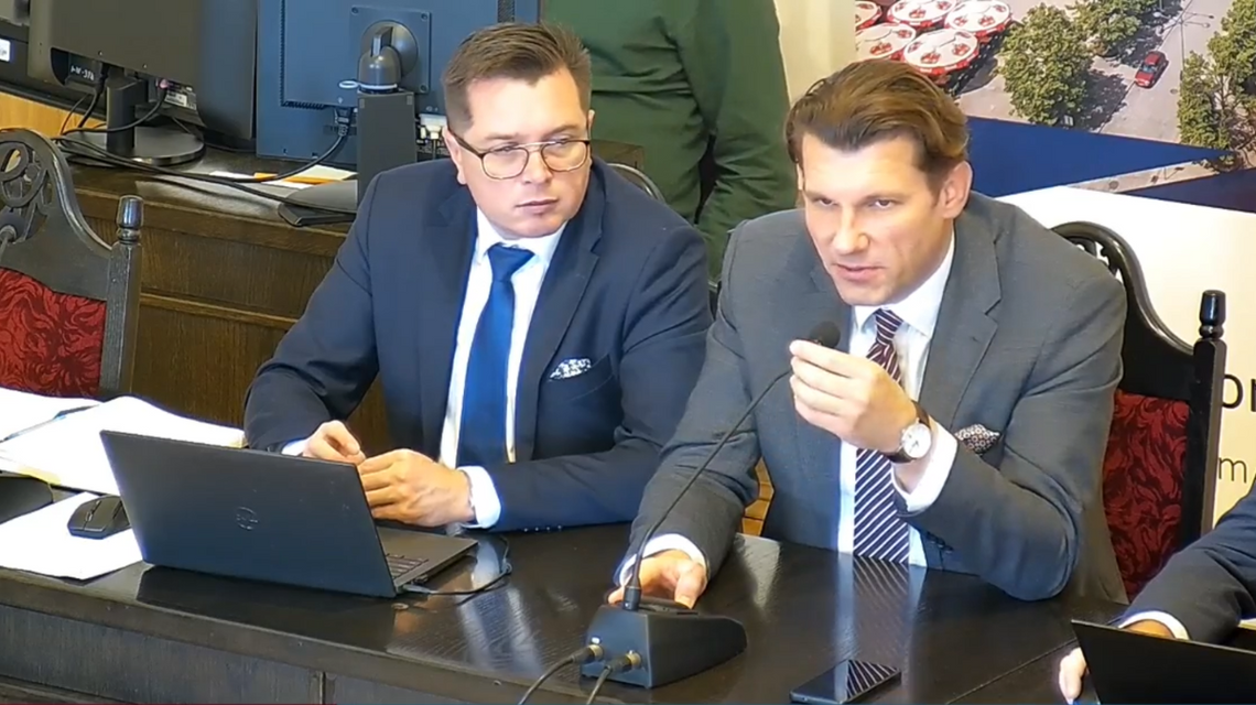 zdjęcie przedstawia dwóch mężczyzn pod krawatem i w garniturach siedzących przy stole
