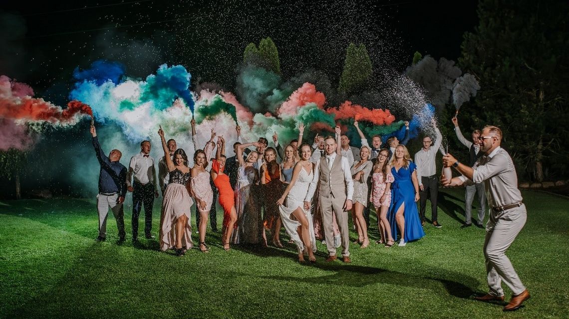 grupa ludzi bawiąca się kolorowymi proszkami podczas przyjęcia weselnego w ogrodzie w świetle ogni sztucznych