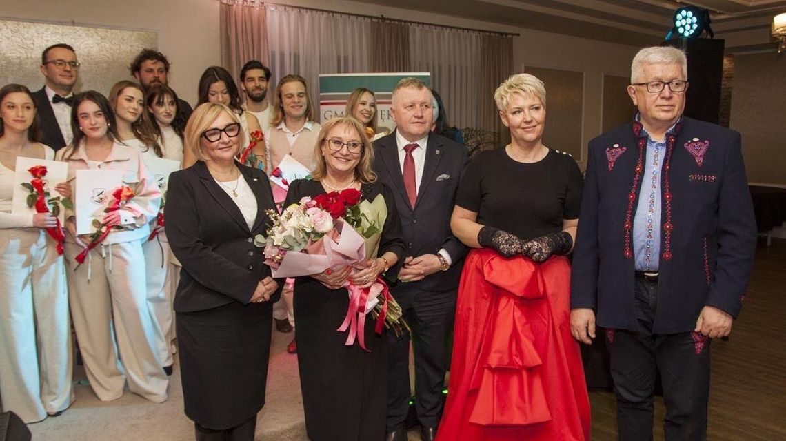 zdjęcie przedstawia kilka osób ubranych w odświętne stroje, w tle młodzież w białych strojach na scenie