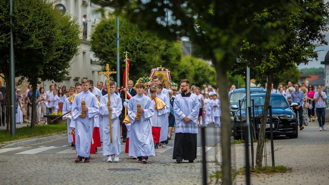 ludzie idący w procesji bożego ciała, ministranci niosą krzyż, idą ulicą miasta, wokoło samochody i drzewa