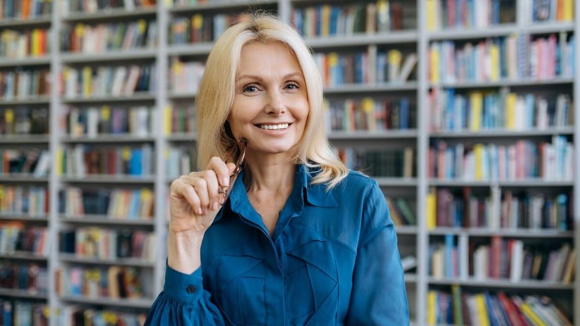 Jak zmienia się praca bibliotekarza i jak się do niej przygotować? zdjęcie przedstawia kobietę z okularami w ręce, w tle półki z kiążkami