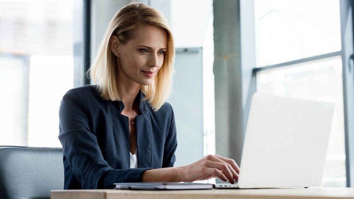 zdjęcie przedstawia kobietę siedzącą przy stole, patrzącą w laptop