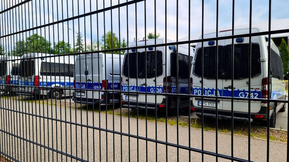 zdjęcie przedstawia kilka radiowozów policji ustawionych w rzędzie, widok przez kratki ogrodzenia