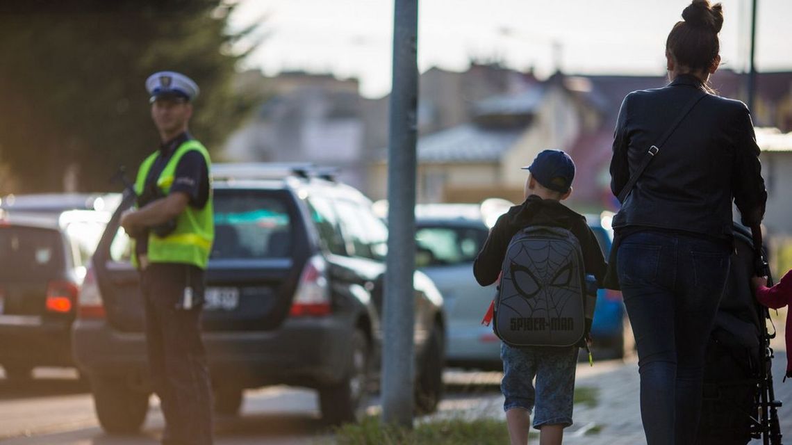 zdjęcie przedstawia dziecko i kobietę pchającą wózek, widok od tył, w tle policjant na ulicy w Gorlicach