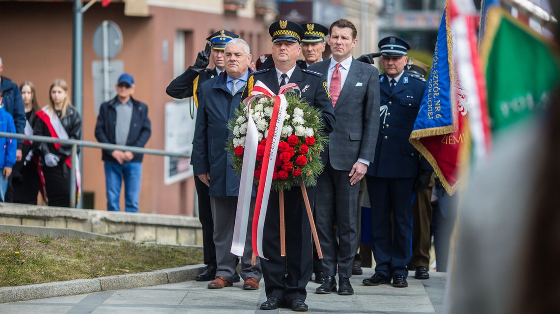 zdjęcie przedstawia grupę osób odświętnie ubranych, pierwsza z nich w mundurze niesie wieniec