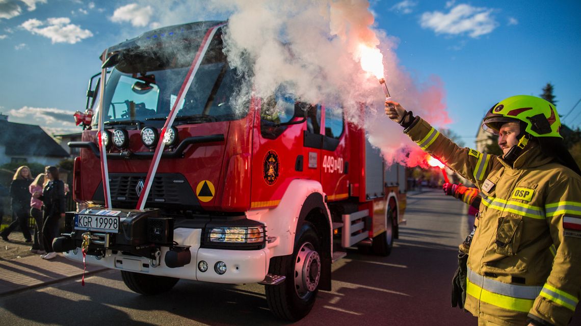 zdjęcie przedstawia druha trzymającego racę, obok samochód strażacki