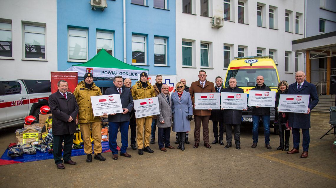 zdjęcie przedstawia grupę osób trzymających tabliczki, za nimi budynek i karetka