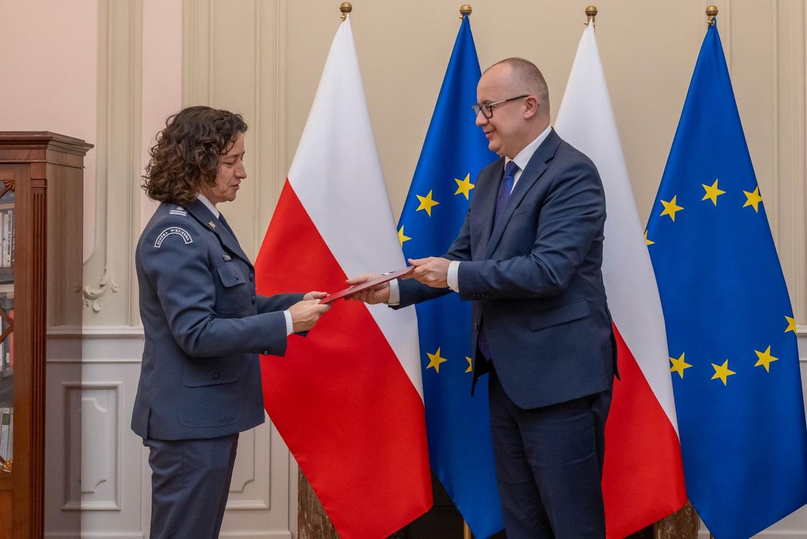 zdjęcie przedstawia kobietę w mundurze, której mężczyzna wręcza teczkę, w tle flagi państwowe