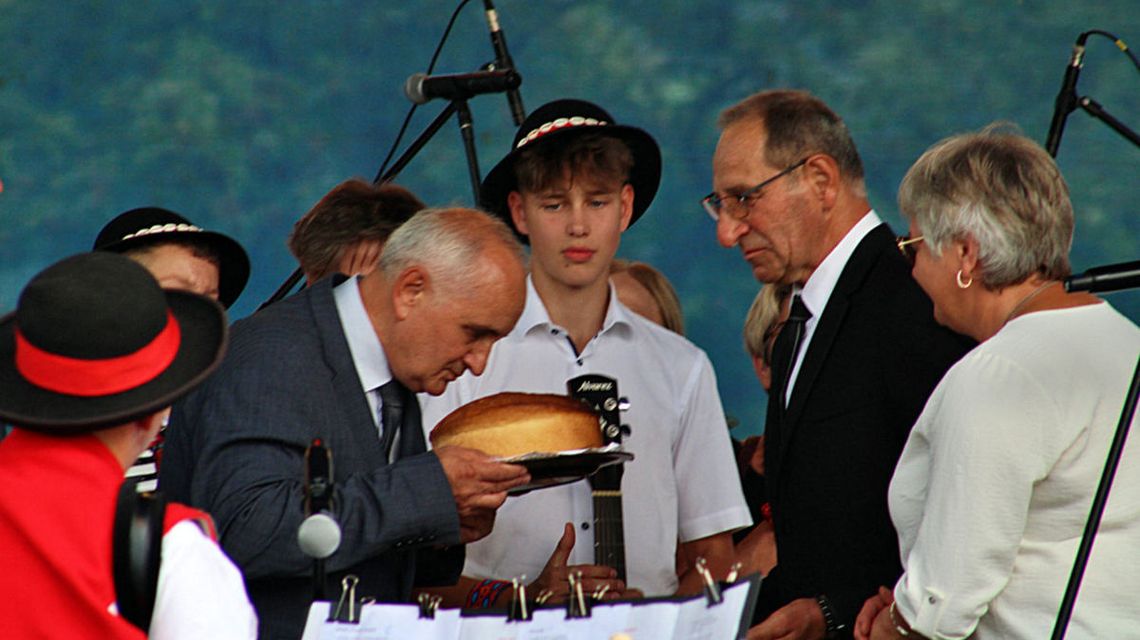 mężczyzna w garniturze pochylający się nad chlebem, żeby go ucałować podczas dożynek, obok inne osoby przyglądają się temu