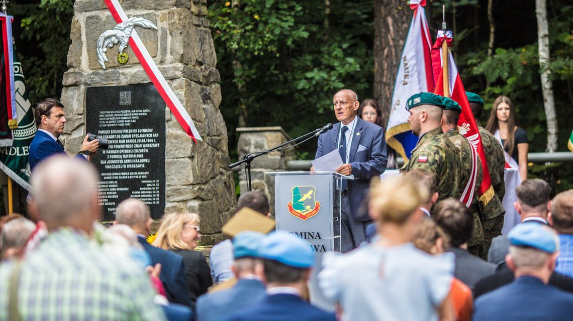 mężczyzna mówiący do mikrofonu przy mównicy przed grupą osób, obok pomnik w krzywej banicy