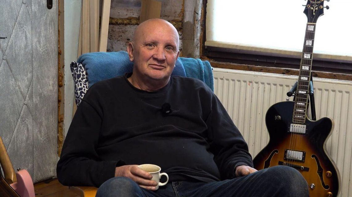 zdjęcie przedstawia mężczyznę z filiżanką w ręce, obok gitara