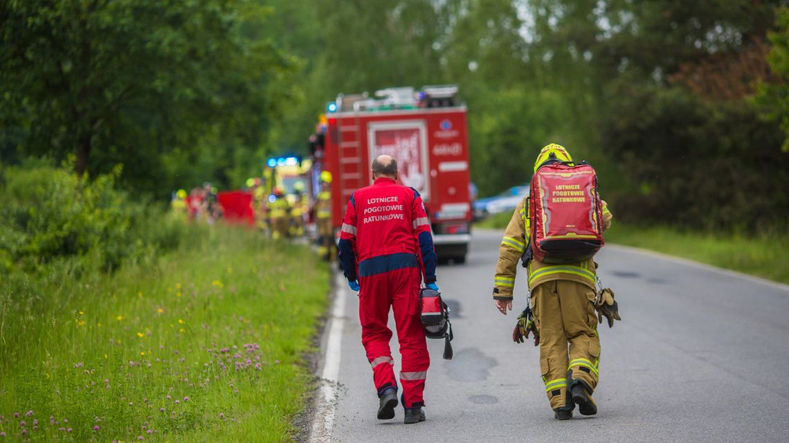 zdjęcie przedstawia dwóch mężczyzn w kombinezonach idących jezdnią, w tle wóz strażacki i karetka