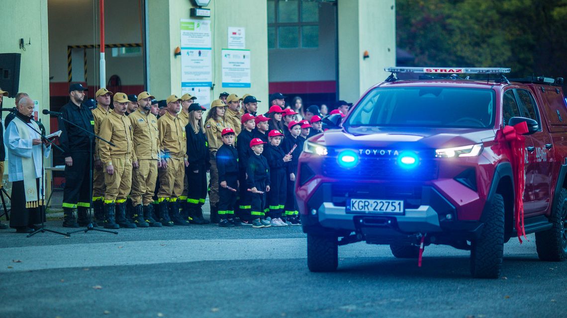 samochód z niebieskimi światłami przed grupą strażaków stojących pod remizą w Szymbarku