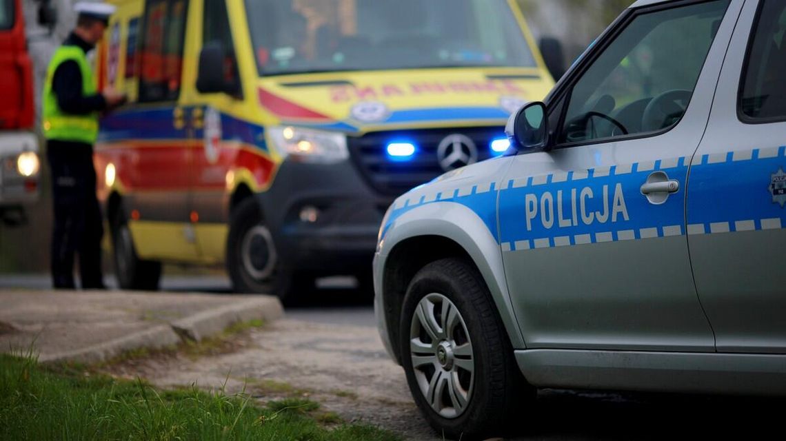 zdjęcie przedstawia policyjny radiowóz na drodze, w tle karetka i policjant