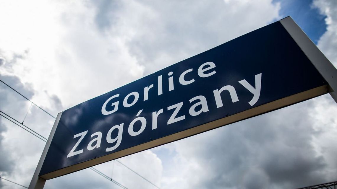 Tablica na stacji PKP Gorlice-Zagórzany w tle zachmurzone niebo i przewody trakcji kolejowej