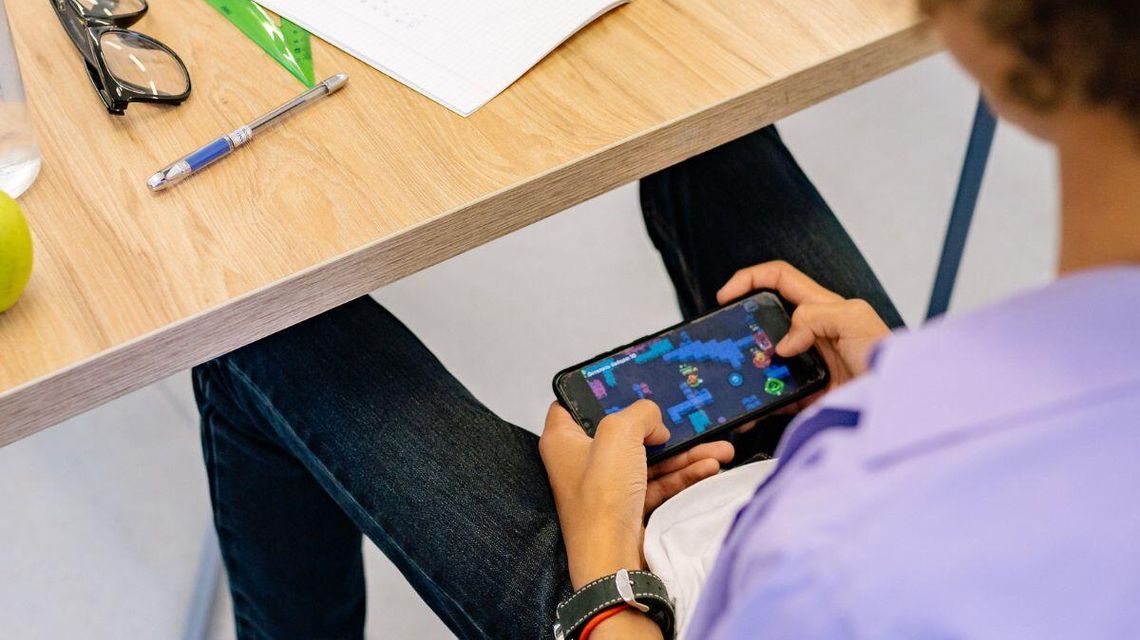 zdjęcie przedstawia telefon komórkowy, który dziecko trzyma siedząc w ławce szkolnej