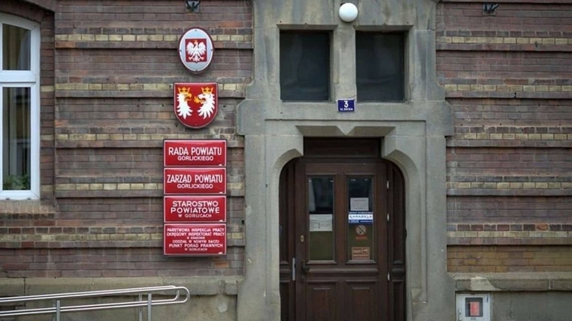 ściana frontowa urzędu, z lewej strony kilka czerwonych tabliczek z napisami o instytucjach obsługujących w bucynku